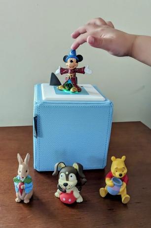 bir çocuğun eli bir mickey mouse oyuncağına uzanıyor ahşap bir masanın üzerinde açık mavi bir tonie kutusu ve bir mickey mouse fantasia tonie