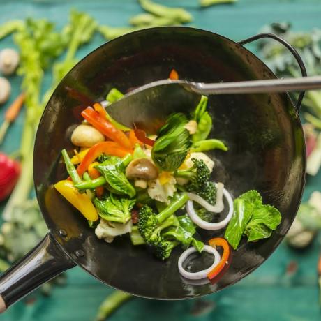 Çeşitli taze renkli pazar sebzelerini sıcak buharlı bir wok'ta karıştırın ve wok'un altındaki turkuaz renkli ahşap masa arka planında sebzelerle soteleyin.